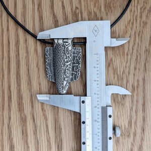 Puede incluir: Colgante plateado con un dise&ntilde;o intrincado y texturizado, suspendido de un cord&oacute;n negro. El colgante se mide con un calibrador met&aacute;lico. El dise&ntilde;o del colgante incluye patrones florales y geom&eacute;tricos. Mide aproximadamente 3.8 cm.