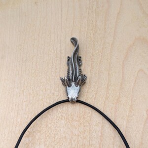 May include: A silver-colored alligator pendant with a black cord necklace. The pendant is detailed with scales and claws. The alligator is facing upwards. The background is a light brown wooden surface.