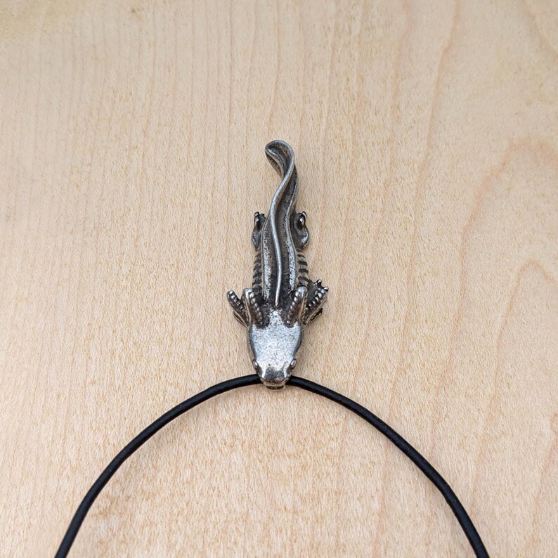 May include: A silver-colored alligator pendant with a black cord necklace. The pendant is detailed with scales and claws. The alligator is facing upwards. The background is a light brown wooden surface.