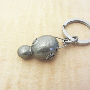 May include: A silver-colored metal keychain featuring a stylized, rounded shape with a smaller, attached sphere. The keychain includes a split ring for attaching keys. The design has a vintage or antique appearance.