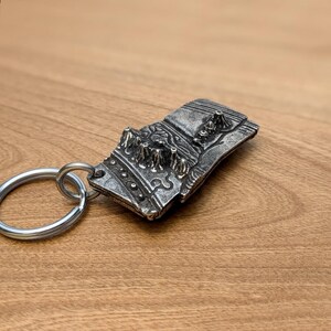 May include: A silver-colored metal keychain featuring a detailed relief design of a mountain range. The rectangular pendant is attached to a silver split ring. The design includes textured peaks and valleys, creating a three-dimensional effect.