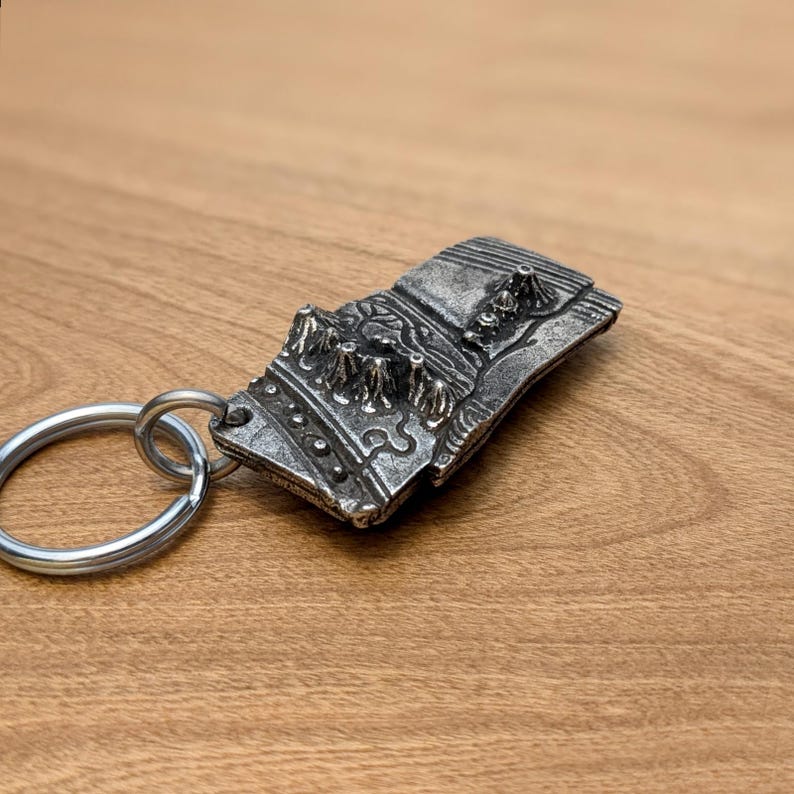 May include: A silver-colored metal keychain featuring a detailed relief design of a mountain range. The rectangular pendant is attached to a silver split ring. The design includes textured peaks and valleys, creating a three-dimensional effect.
