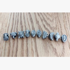 May include: A collection of ten individual silver-toned metal beads arranged in a horizontal row. Each bead features a unique embossed texture, including geometric patterns, dotted surfaces, and linear grooves, with a central hole for threading.