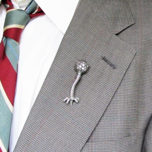 May include: A silver-toned bacteriophage lapel pin with a round head and a long, thin body with legs. The pin is attached to a gray suit jacket.
