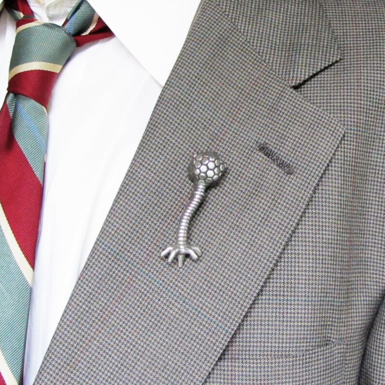 May include: A silver-toned bacteriophage lapel pin with a round head and a long, thin body with legs. The pin is attached to a gray suit jacket.