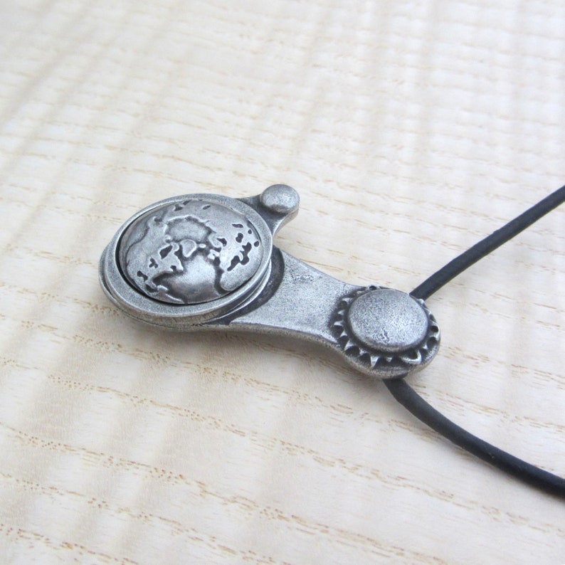 May include: A silver pendant necklace with a globe design. The pendant is attached to a black cord.