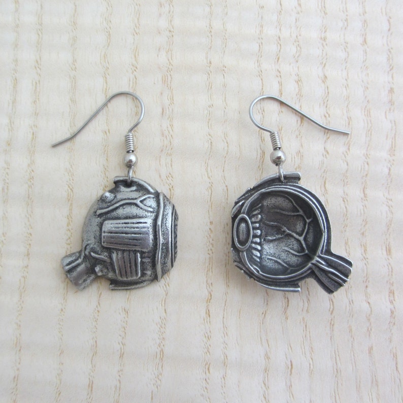 May include: A pair of silver earrings shaped like a human eye. The earrings are detailed with the internal structures of the eye, including the iris, pupil, and lens.