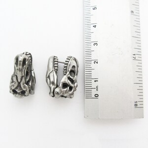 May include: Two silver-colored earrings shaped like dinosaur skulls. The earrings are detailed with textured surfaces and open spaces, and are approximately 1 inch in length. The earrings are displayed on a white background next to a ruler.