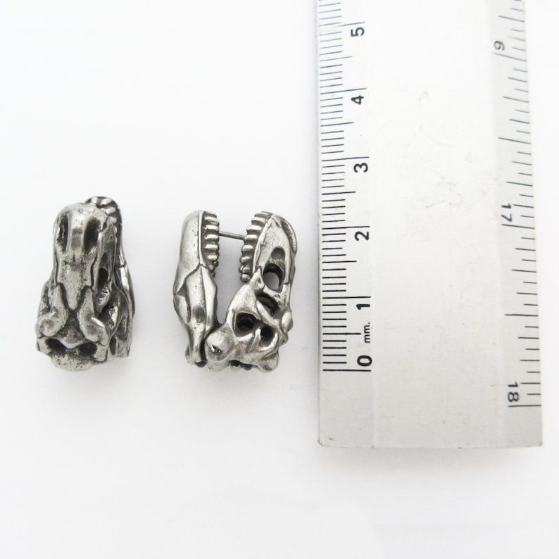 May include: Two silver-colored earrings shaped like dinosaur skulls. The earrings are detailed with textured surfaces and open spaces, and are approximately 1 inch in length. The earrings are displayed on a white background next to a ruler.