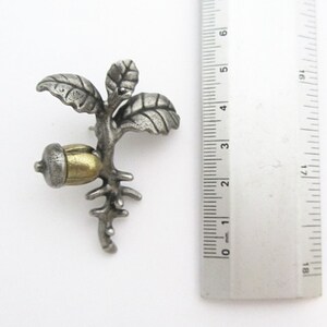 May include: A silver-toned metal brooch in the shape of a sprouting acorn with leaves. The acorn has a gold-colored cap. The brooch is approximately 1.5 inches in height and is set against a white background.