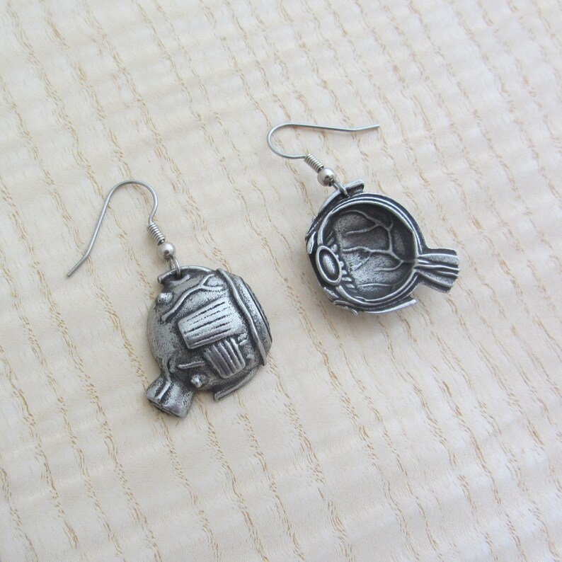 May include: A pair of silver earrings shaped like a human eye. The earrings are detailed with the internal anatomy of the eye, including the iris, pupil, and optic nerve.