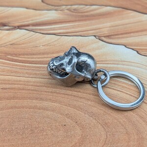 Puede incluir: Un llavero de metal plateado con un dije tridimensional detallado de un cr&aacute;neo de primate. El cr&aacute;neo est&aacute; unido a una anilla met&aacute;lica circular y muestra caracter&iacute;sticas anat&oacute;micas realistas, incluyendo cuencas oculares, mand&iacute;bula y dientes.