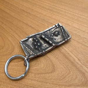 May include: A silver-toned metal keychain with a rectangular, textured pendant. The pendant has an intricate design with raised details and is attached to a silver key ring. The keychain rests on a wooden surface.