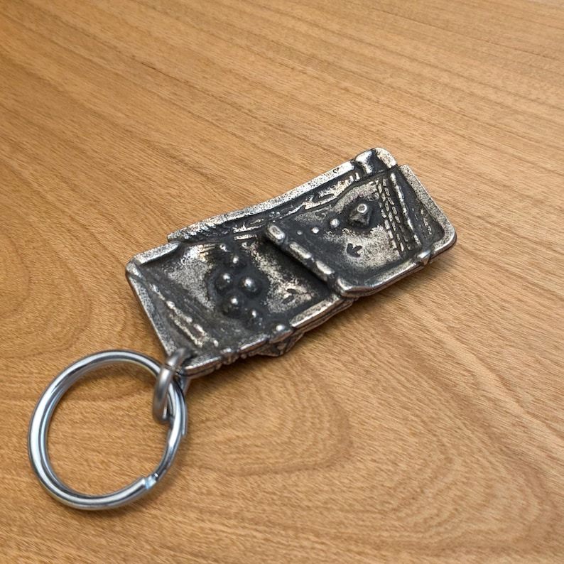 May include: A silver-toned metal keychain with a rectangular, textured pendant. The pendant has an intricate design with raised details and is attached to a silver key ring. The keychain rests on a wooden surface.