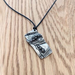 Op de afbeelding: Een zilverkleurige hangerketting met een zwart koord. De hanger heeft een abstract ontwerp met ge&euml;tste details en een getextureerd oppervlak. De ketting wordt getoond op een houten oppervlak.
