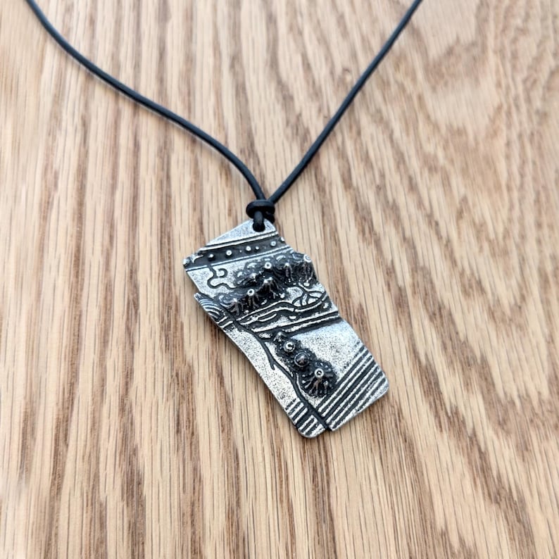 Op de afbeelding: Een zilverkleurige hangerketting met een zwart koord. De hanger heeft een abstract ontwerp met ge&euml;tste details en een getextureerd oppervlak. De ketting wordt getoond op een houten oppervlak.