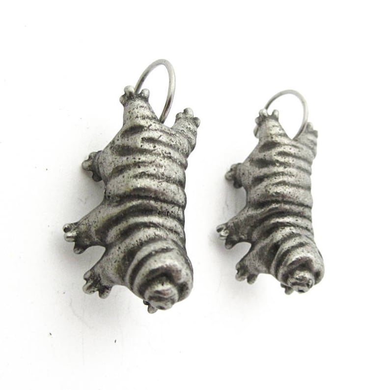 May include: A pair of silver-toned metal pendants shaped like tardigrades, also known as water bears. Each pendant features a segmented, textured body with eight distinct legs and a small circular loop at the top for attachment to a chain or cord.