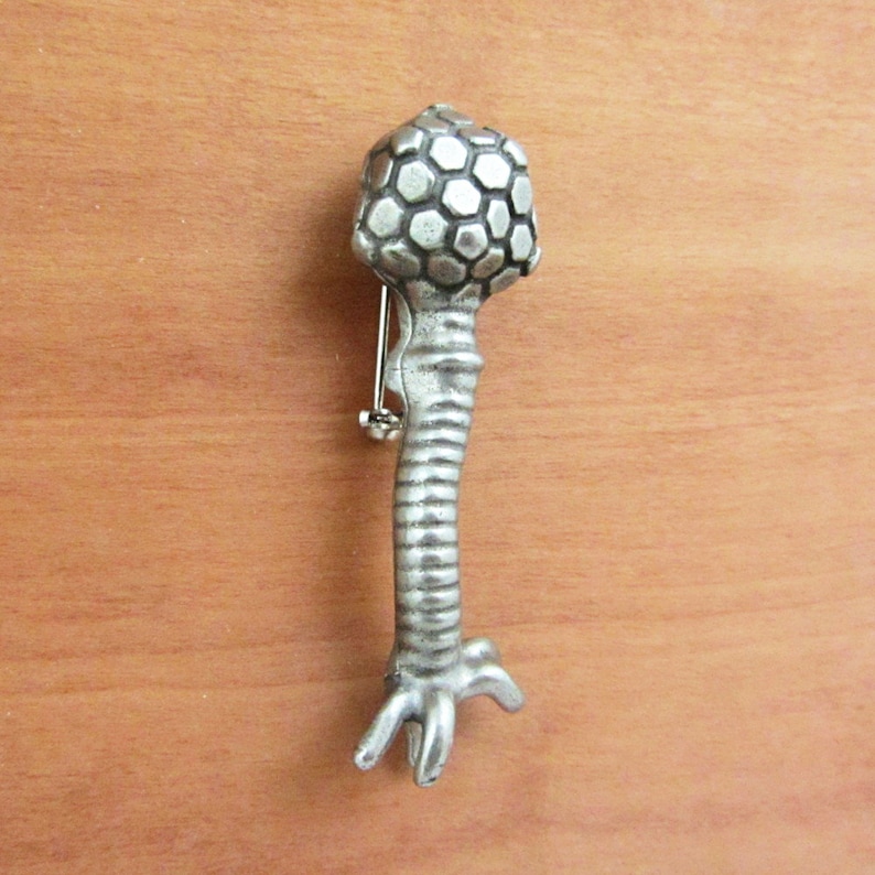 May include: A silver-toned brooch in the shape of a stylized chicken leg. The leg is textured and has a three-toed foot. The top of the leg is a round, textured ball.