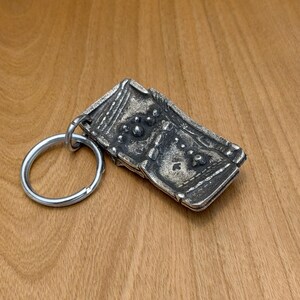 May include: A rectangular, silver-toned metal keychain with a detailed, textured design. The keychain features a large, round key ring and an ornate, rectangular pendant with raised patterns and small, circular details. The background is a wooden surface.