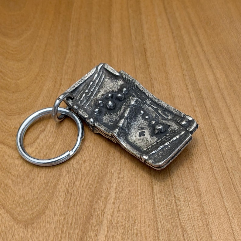 May include: A rectangular, silver-toned metal keychain with a detailed, textured design. The keychain features a large, round key ring and an ornate, rectangular pendant with raised patterns and small, circular details. The background is a wooden surface.