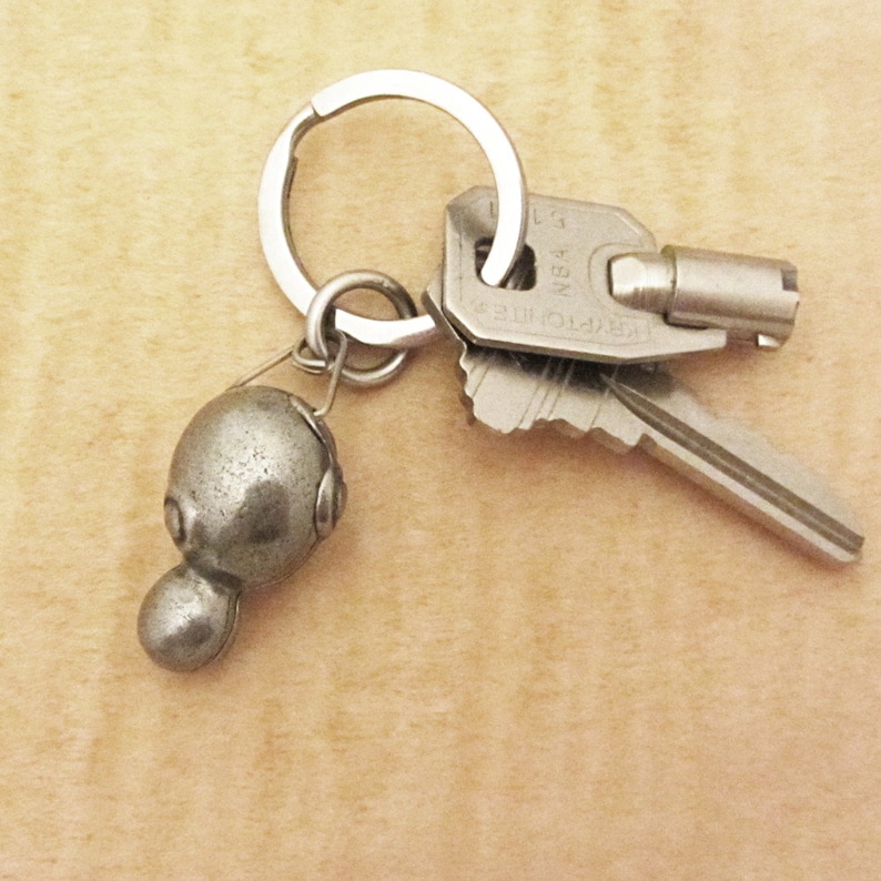 May include: A silver-colored keychain with a small, rounded charm and two keys. The charm has a unique, abstract design. The keys are attached to a split ring, and the keychain is on a light-colored surface.
