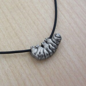 May include: A silver-colored caterpillar pendant on a black cord necklace. The caterpillar has detailed ridges and a curled head. The necklace is displayed on a light brown wooden surface.