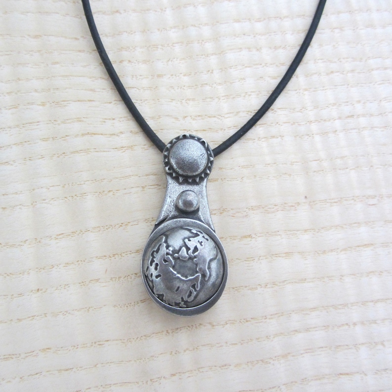 May include: A silver necklace with a pendant featuring a detailed globe design. The pendant has a textured surface and a small circle on the top.