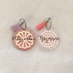 May include: Two pink and white glitter resin keychains with silver key rings and pink and purple tassels. The keychains have a white and pink floral design and the names "Alessandra" and "Yasmerie" written in black.