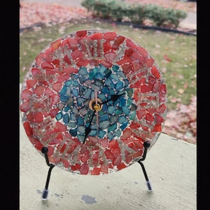 Puede incluir: Un reloj de mosaico con piezas de vidrio rojo, azul y blanco. El reloj está en un soporte de metal negro.