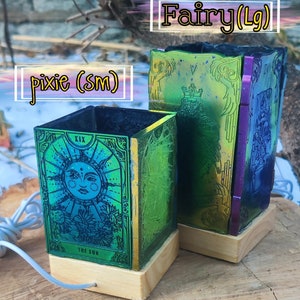 May include: Two iridescent glass lamps with wooden bases. The smaller lamp features a depiction of the sun from a tarot deck, while the larger lamp has a design of a king from a tarot deck. The lamps are labelled "pixie (sm)" and "fairy (Lg)".
