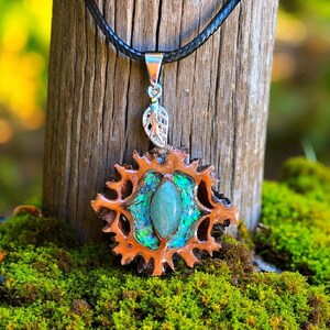 Pode incluir: Um colar com pingente artesanal com um design único. O pingente tem uma moldura externa marrom e pontiaguda com uma pedra central ovalada verde claro. A pedra é cercada por detalhes iridescentes azuis e verdes. O colar pende de um cordão de couro preto.