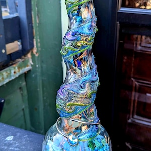 May include: A decorative glass bottle with a swirling, colorful design. The bottle is adorned with blue, green, and gold accents, and sits on a wooden base. The base of the bottle is filled with blue and green stones.