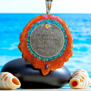 May include: A pendant with a silver-colored circular charm that reads "Blessed are the Curious, for they shall have adventures." The pendant has an orange and blue decorative border and a silver bail. The pendant is resting on a black stone with seashells in the foreground.