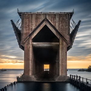May include: A large, weathered concrete structure with a tunnel-like opening, standing on a body of water. The structure has a unique, angular design with a pointed roof and a series of vertical supports. The water is calm and reflects the sky, which is a mix of blue and orange hues. The structure appears to be an old industrial building, possibly a grain elevator or a dock.