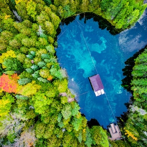 May include: An aerial view of a clear blue spring surrounded by a lush green forest with some fall foliage. A small wooden dock is in the water.