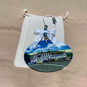 Op de afbeelding: Ovale kerstversiering met een vintage afbeelding van het Grand View Ship Hotel, blauwe lucht en auto's. Het is versierd met een witte strik, blauwe kralen en een ophangkoord. De versiering wordt gepresenteerd met een klein, off-white trekkoordzakje.