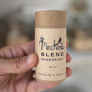 K&ouml;nnte beinhalten: Zylindrischer Deodorantbeh&auml;lter aus braunem Karton. Das Etikett zeigt das Wort "Merchants" in einer stilisierten Schriftart mit einer Vogelgrafik. Der Text "BLEND DEODORANT" und "2+ oz" sind ebenfalls sichtbar. Das Produkt wird gehalten.