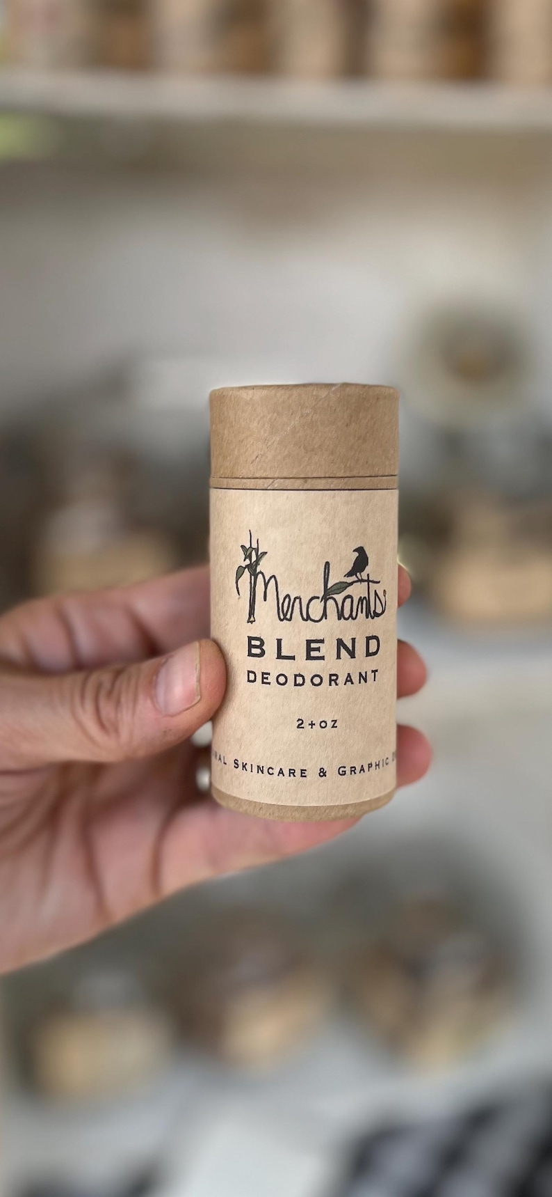 K&ouml;nnte beinhalten: Zylindrischer Deodorantbeh&auml;lter aus braunem Karton. Das Etikett zeigt das Wort "Merchants" in einer stilisierten Schriftart mit einer Vogelgrafik. Der Text "BLEND DEODORANT" und "2+ oz" sind ebenfalls sichtbar. Das Produkt wird gehalten.