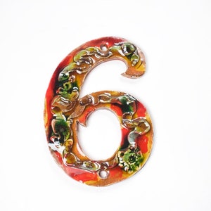 May include: A ceramic house number six with a colorful, floral design. The number is painted in shades of red, orange, yellow, green, and brown.