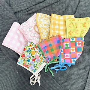 May include: A collection of colorful bonnets with various patterns, including floral, gingham, and geometric designs. The bonnets feature pastel pinks, yellows, and blues, with tie closures. The bonnets are made of soft fabric.