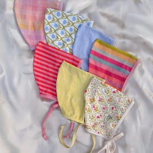 May include: A collection of seven fabric bonnets featuring various patterns and colors, including pink plaid, blue floral, solid light blue, red stripes, multicolored stripes, solid yellow, and a white floral print with pink and green accents.