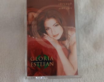 May include: A cassette tape featuring the album Christmas Through Your Eyes by Gloria Estefan. The cover art displays a portrait of the artist against a deep red background with gold text and a large letter C overlaying the image.