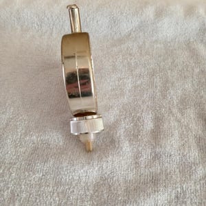 Pu&ograve; includere: Un tappo per bottiglia di vino in metallo argentato con un corpo a forma di anello e una parte superiore cilindrica. Il tappo ha una base filettata scanalata e un piccolo beccuccio conico. L'oggetto &egrave; su una superficie bianca e strutturata.