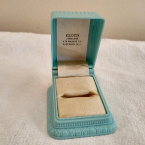 May include: An open, vintage, light blue jewelry box with a beige interior. The box is rectangular with a hinged lid and a decorative base. The text "ELLIOTT'S JEWELERS 122 MARKET ST. PATERSON, N. J." is printed inside the lid.