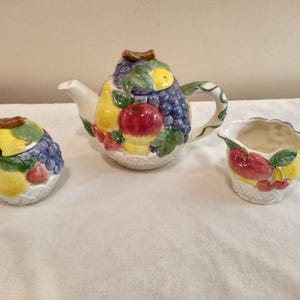May include: A ceramic tea set featuring a teapot, sugar bowl, and creamer. The teapot is adorned with a fruit design, including grapes, apples, and pears. The sugar bowl and creamer also feature fruit motifs. The set has a white base with a woven basket design.