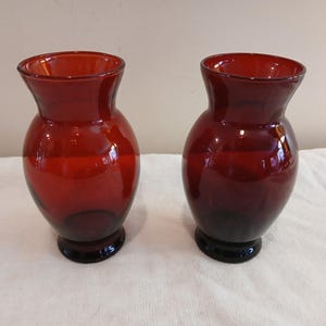 May include: A pair of matching deep red glass vases with a bulbous body, a flared rim, and a circular pedestal base. The glass is translucent, allowing light to pass through the rich crimson color, creating subtle highlights on the curved surfaces.
