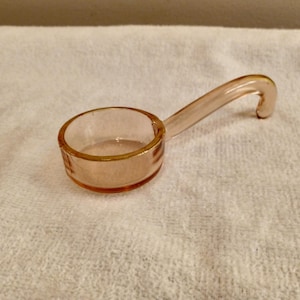May include: A small, light pink glass measuring spoon with a curved handle. The spoon has a round bowl and is made of transparent glass. The handle curves upward, ending in a small hook. The spoon is resting on a white, textured surface.