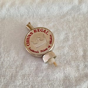 Pu&ograve; includere: Un tappo per bottiglia Chivas Regal argentato con scritte rosse e un'emblema a corona. Il tappo ha un piccolo beccuccio e un tappo a vite. Il testo recita "Chivas Regal Scotland's Princes of Whiskies."