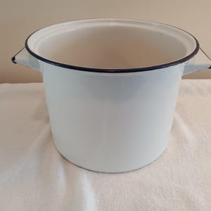 May include: A white enamel pot with a dark blue rim and handles. The pot is cylindrical with a wide opening and is sitting on a white surface. The pot is empty.