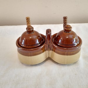 May include: A ceramic condiment server with two connected compartments. Each compartment has a brown lid with a wooden handle. The base is a light beige color, with a brown band around the top. A handle connects the two compartments.